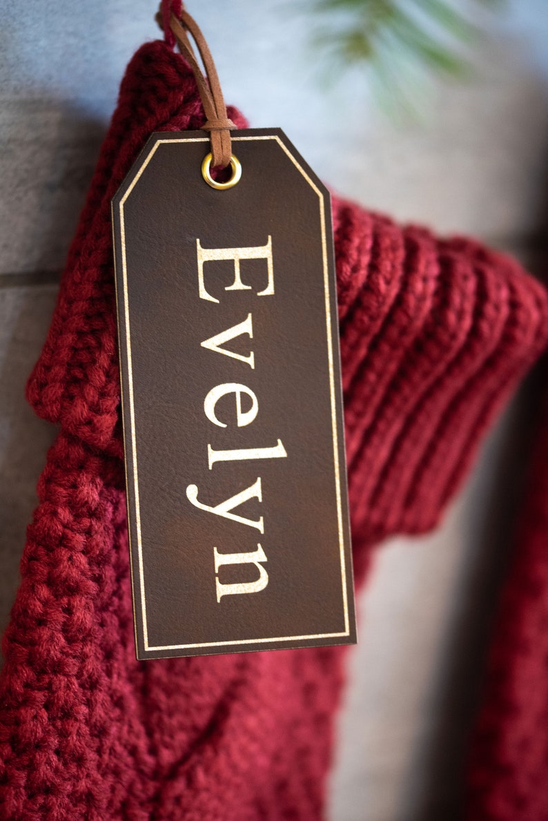 Op de afbeelding: Een rode gebreide sok met een bruin leren label waarop "Evelyn" in gouden letters staat.