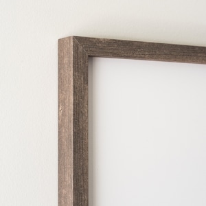 May include: Close-up of a rectangular picture frame with a weathered, dark brown wood grain finish. The frame has a simple, classic design and is set against a white wall. The corner of the frame is visible.