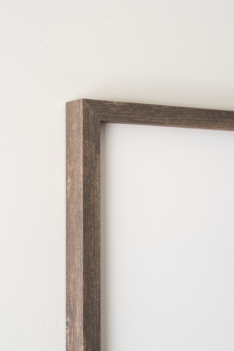 May include: Close-up of a rectangular picture frame with a weathered, dark brown wood grain finish. The frame has a simple, classic design and is set against a white wall. The corner of the frame is visible.