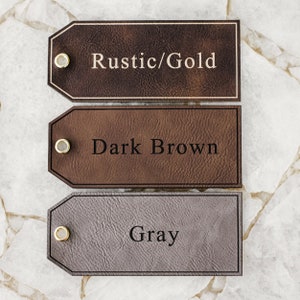 Op de afbeelding: Drie leren labels met verschillende kleuren en tekst. Het bovenste label is bruin met de tekst "Rustic/Gold". Het middelste label is donkerbruin met de tekst "Dark Brown". Het onderste label is grijs met de tekst "Gray".