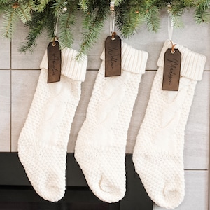 Op de afbeelding: Drie witte gebreide kousen met bruine leren naamplaatjes. De naamplaatjes zijn gepersonaliseerd met de namen "Amelia", "Willow" en "Madison".