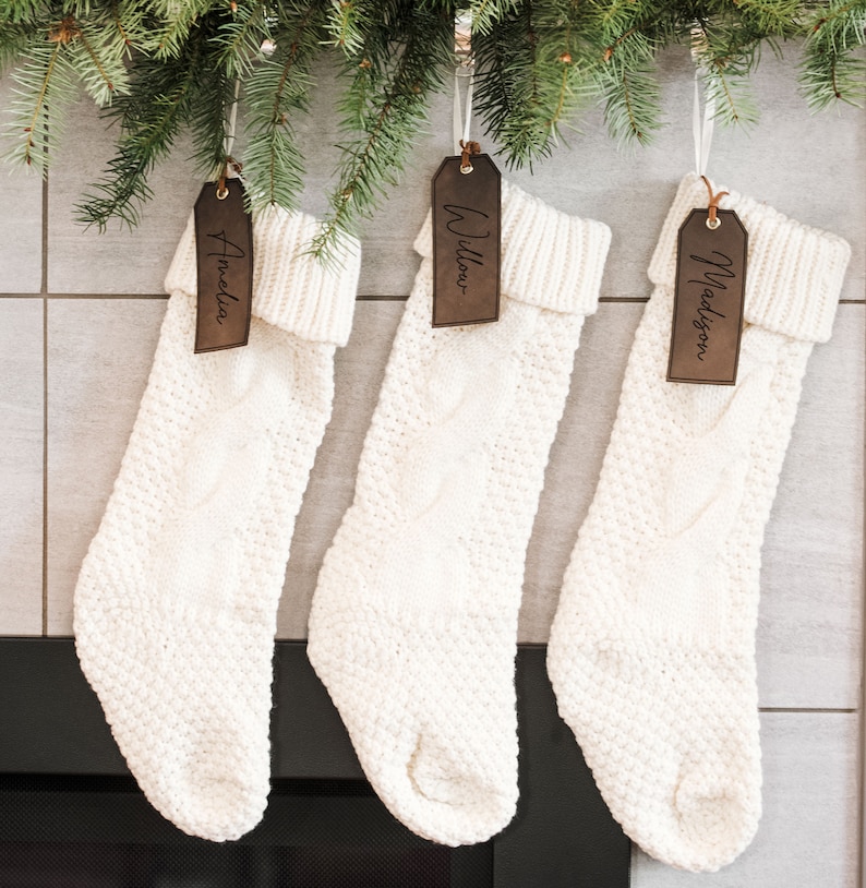 Op de afbeelding: Drie witte gebreide kousen met bruine leren naamplaatjes. De naamplaatjes zijn gepersonaliseerd met de namen "Amelia", "Willow" en "Madison".