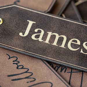 Op de afbeelding: Een bruin lederen cadeau-etiket met een gouden metalen oogje en de naam "James" gedrukt in gouden letters.