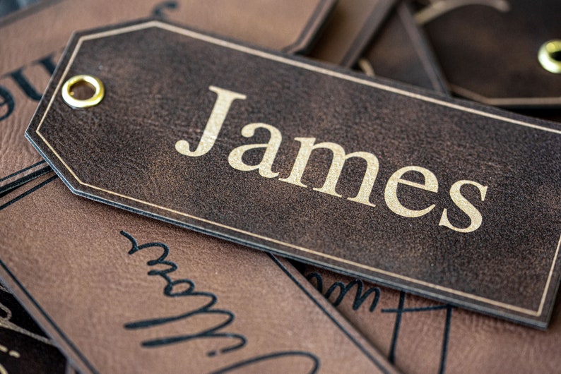 Op de afbeelding: Een bruin lederen cadeau-etiket met een gouden metalen oogje en de naam "James" gedrukt in gouden letters.