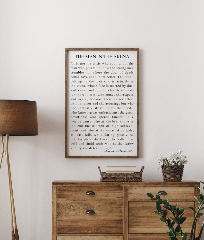 May include: A framed print with the text "THE MAN IN THE ARENA" and a quote by Theodore Roosevelt. The print is in a dark brown frame and is displayed on a wooden dresser. A lamp and decorative items are also visible.