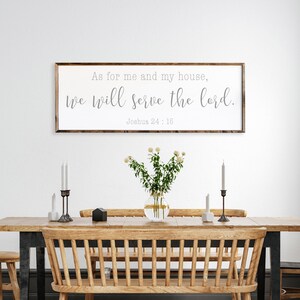 May include: A framed wood sign with the text "As for me and my house, we will serve the Lord. Joshua 24:15" hanging on a white wall above a wooden dining table with two chairs and a vase of white flowers.