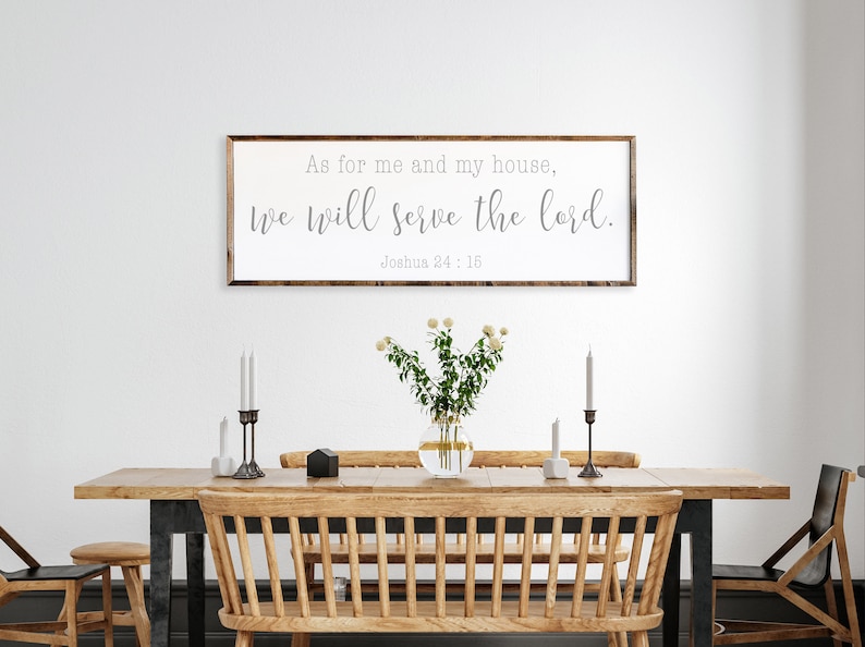 May include: A framed wood sign with the text "As for me and my house, we will serve the Lord. Joshua 24:15" hanging on a white wall above a wooden dining table with two chairs and a vase of white flowers.