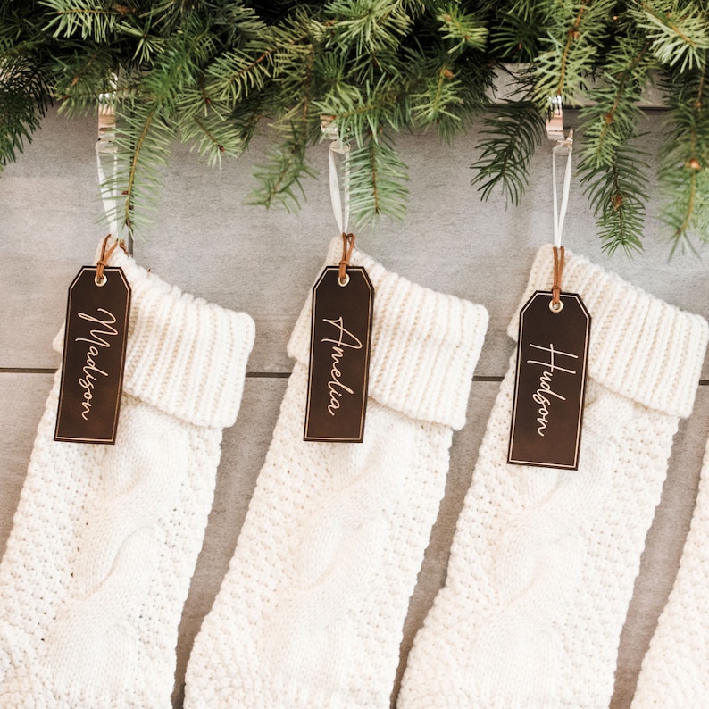 Op de afbeelding: Drie witte gebreide kousen met bruine leren labels die aan een groene krans hangen. De labels zijn gepersonaliseerd met de namen "Madison", "Amelia" en "Hudson".