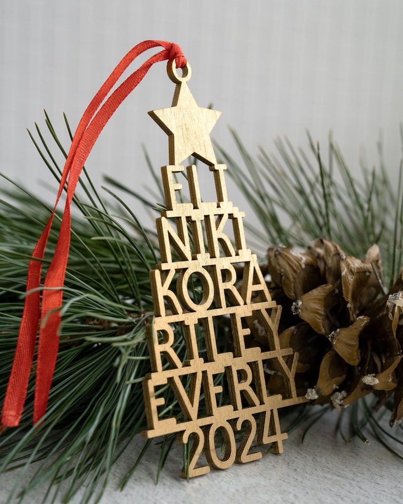 May include: Gold wooden Christmas tree ornament with the names Eli, Niki, Kora, Riley, Everly and the year 2024.