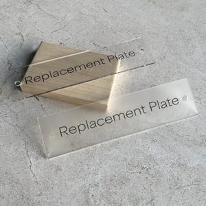 May include: Two clear acrylic replacement plates with the text "Replacement Plate" printed in black. One plate is slightly frosted, and the other is transparent. The plates are resting on a light-colored wooden block.