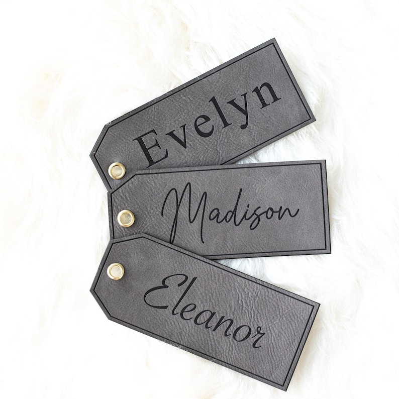 Op de afbeelding: Drie grijze leren cadeau-etiketten met zwarte letters. De etiketten liggen op elkaar gestapeld. Het bovenste etiket zegt "Evelyn", het middelste etiket zegt "Madison" en het onderste etiket zegt "Eleanor".