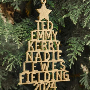 May include: Gold wooden Christmas tree ornament with the names Ted, Emmy, Kerry, Nadia, Lewis, Fielding and the year 2024.