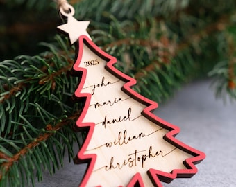 May include: A wooden Christmas tree ornament with a red outline and a star at the top. The ornament has the year 2025 and names engraved on it. It hangs from a twine loop, set against a backdrop of evergreen branches.