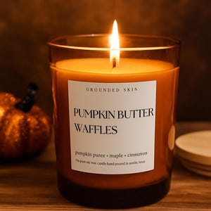 Op de afbeelding: Een brandende oranje kaars in een heldere glazen pot met een wit etiket met de tekst "PUMPKIN BUTTER WAFFLES". De kaars is geparfumeerd met pompoenpuree, esdoorn en kaneel.