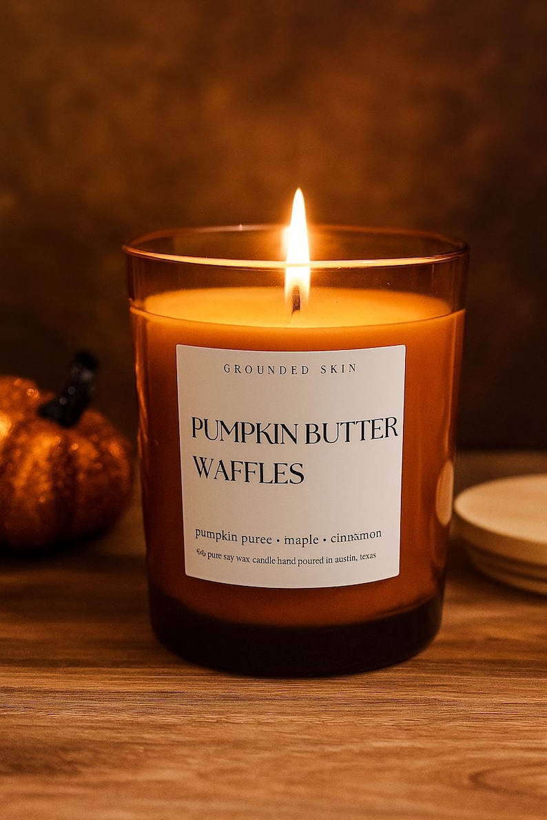 Op de afbeelding: Een brandende oranje kaars in een heldere glazen pot met een wit etiket met de tekst "PUMPKIN BUTTER WAFFLES". De kaars is geparfumeerd met pompoenpuree, esdoorn en kaneel.