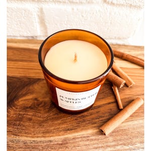 Op de afbeelding: Een kaars in een bruine glazen pot met een wit etiket met de tekst "Pumpkin Butter Waffles". De kaars staat op een houten oppervlak met verschillende kaneelstokjes. De kaars heeft een witte was en een enkele lont.