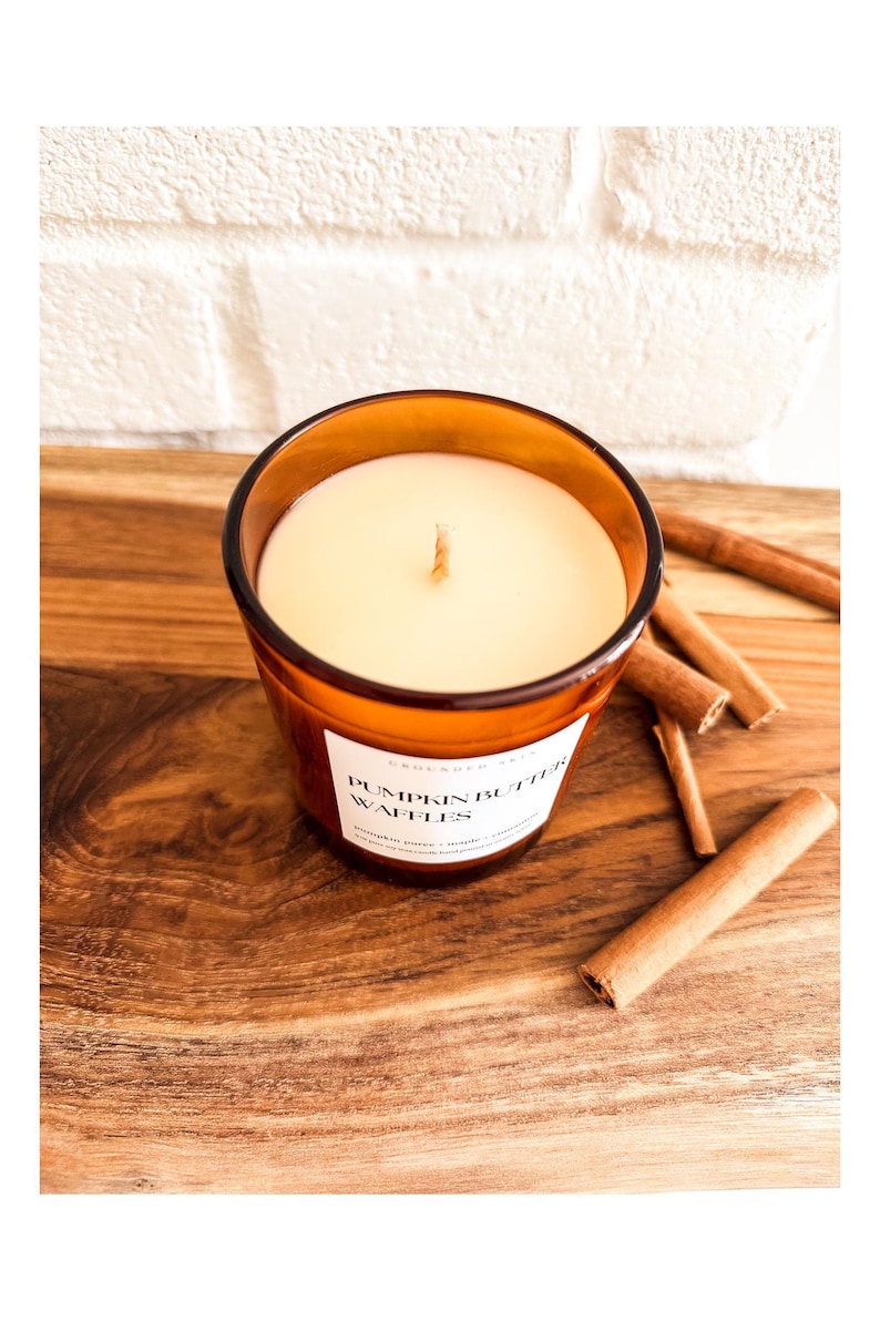 Op de afbeelding: Een kaars in een bruine glazen pot met een wit etiket met de tekst "Pumpkin Butter Waffles". De kaars staat op een houten oppervlak met verschillende kaneelstokjes. De kaars heeft een witte was en een enkele lont.