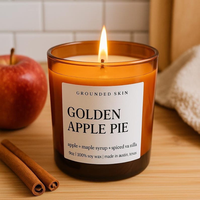 Op de afbeelding: Een brandende kaars in een glazen pot met de tekst "GOLDEN APPLE PIE". De kaars staat naast een rode appel en twee kaneelstokjes. Het etiket luidt "apple + maple syrup + spiced vanilla" en "9oz | 100% soy wax".