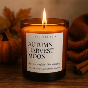 Op de afbeelding: Een brandende kaars in een glazen pot met de tekst "AUTUMN HARVEST MOON". De kaars is oranje met een wit etiket met de tekst "GROUNDED SKIN". De geur is vijg, warme kruiden en bloedsinaasappel. De kaars is 255g en gemaakt in Austin, Texas.
