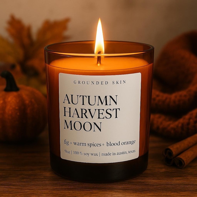 Op de afbeelding: Een brandende kaars in een glazen pot met de tekst "AUTUMN HARVEST MOON". De kaars is oranje met een wit etiket met de tekst "GROUNDED SKIN". De geur is vijg, warme kruiden en bloedsinaasappel. De kaars is 255g en gemaakt in Austin, Texas.