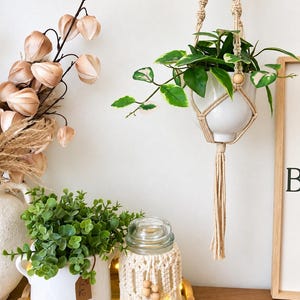May include: A white ceramic plant pot held by a beige macrame hanger with wooden beads and a long tassel. The hanger supports a green and white variegated plant. Nearby, a white pot with green foliage and a macrame-covered glass jar are visible.