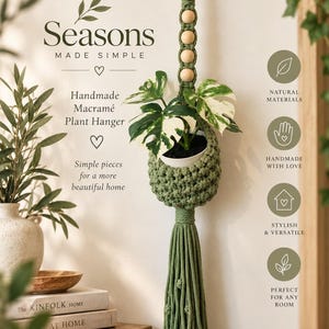 May include: A sage green macramé plant hanger featuring a wooden ring at the top, four light-coloured wooden beads, and a long tasseled bottom. The hanger holds a white pot containing a variegated monstera plant with green and white leaves.