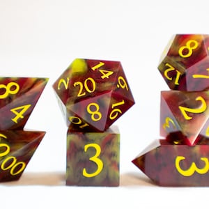 May include: A set of seven polyhedral dice with a red, black, and yellow marbled pattern. The dice are numbered with yellow numbers. The dice are arranged in a row on a white surface.