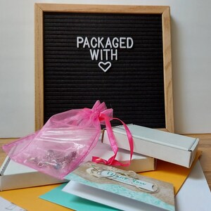 May include: A pink organza bag filled with small items, a white box with a thank you card, and a black letter board with the text "PACKAGED WITH" and a white heart.