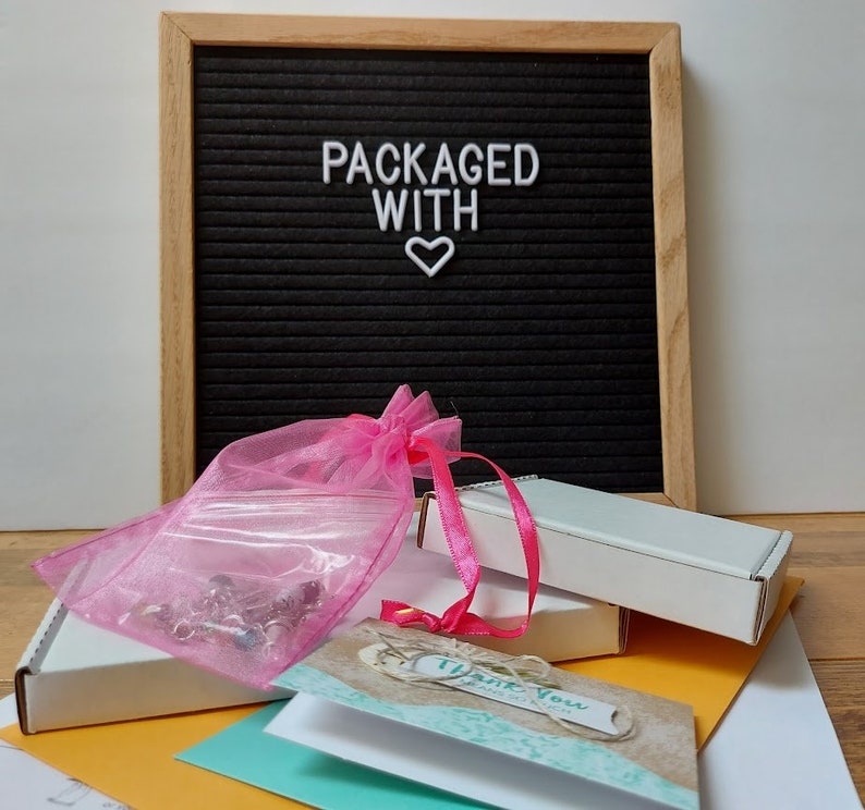 May include: A pink organza bag filled with small items, a white box with a thank you card, and a black letter board with the text "PACKAGED WITH" and a white heart.