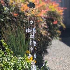 May include: A decorative wind chime featuring a black cloud-shaped top and multiple strands of clear, faceted crystal beads and spheres. The wind chime hangs outdoors with a blurred background of green and orange foliage.
