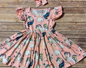 May include: A pink dress with a unicorn pattern, white clouds, and stars. The dress has short sleeves and a flared skirt. A matching pink bow is also present. The dress is made of a soft fabric and is suitable for children.