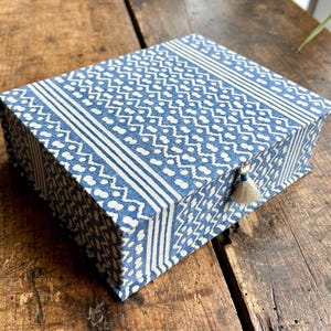 May include: A rectangular storage box covered in blue and white fabric featuring a repeating geometric pattern of zigzags and dots. The lid is secured with a small, off-white tassel attached to the front edge, creating a simple closure mechanism.
