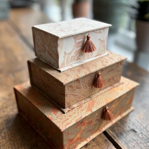 May include: A stack of three rectangular boxes with lids, arranged from smallest on top to largest on the bottom. Each box features a patterned surface in shades of cream, tan, and burnt orange, and includes a small, dangling brown tassel.