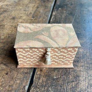 May include: A rectangular fabric-covered box featuring a lid with a large-scale floral pattern in muted peach and olive tones. The front panel displays a repeating wavy line and dot pattern in similar colors, accented by a small cream-colored tassel.