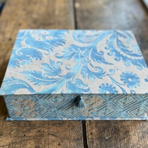 May include: A rectangular storage box covered in fabric featuring a light blue and beige floral paisley pattern on the lid. The front panel displays a blue and beige geometric diamond design with a small blue tassel attached to the center latch.