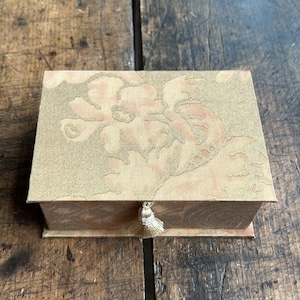 May include: A rectangular box covered in a light tan fabric featuring a subtle, tonal floral pattern. The lid is hinged and includes a small, decorative tassel hanging from the center front edge, providing a simple closure mechanism for the container.