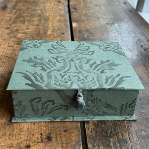 May include: A rectangular sage green box with a dark green damask pattern printed on the lid and sides. The box features a small, matching sage green fabric tassel attached to the center front edge, serving as a decorative pull.
