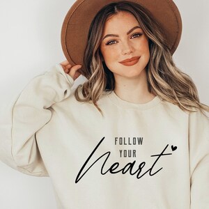 Op de afbeelding: Crèmekleurige sweatshirt met de tekst "Follow Your Heart" in een zwart script. Een klein zwart hartje staat naast het woord "Heart". De sweatshirt wordt gecombineerd met een blauwe jeans en een bruine hoed.