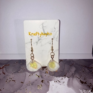 Puede incluir: Pendientes de concha con ganchos y detalles dorados. Las conchas blancas tienen un interior amarillo y brillante. Los pendientes se muestran en una tarjeta blanca con el texto "Kraftykayko".