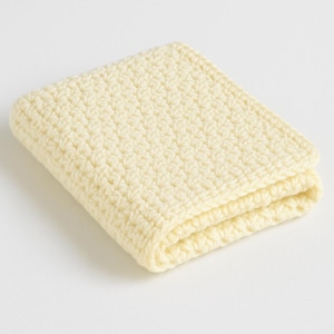 Op de afbeelding: Gevouwen, vierkante, crèmekleurige gehaakte washand. De handgemaakte doek is dubbelgevouwen en toont het gestructureerde steekpatroon. De zachte, neutrale kleur en het eenvoudige ontwerp suggereren een focus op natuurlijke materialen en woondecoratie.