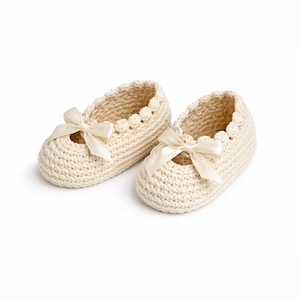 Op de afbeelding: Een paar crèmekleurige, gehaakte babyschoentjes. Elke schoen heeft een delicate geschulpte rand en een satijnen strik. De schoenen zijn ontworpen in een Mary Jane-stijl, perfect voor een pasgeborene of baby.