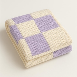 Può includere: Coperta all'uncinetto piegata con un motivo a scacchiera. La coperta è realizzata con filato color crema e lavanda. I quadrati sono di dimensioni uniformi e la coperta è ben piegata.