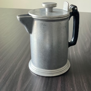 May include: A small, silver-colored French press coffee maker with a black handle. The press has a cylindrical body, a lid with a small knob, and a spout for pouring. The base has a decorative, tiered design.