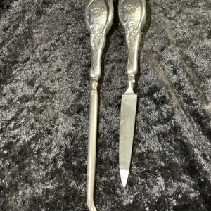 以下が含まれることがあります： 銀色の装飾的なツールが2つセットになっています。ツールは花柄のデザインで、フックとファイルの形をしています。