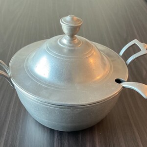May include: A pewter soup tureen with a lid and two handles. The tureen has a round body and a small knob on top of the lid. A spoon rests on the side of the tureen. The tureen is on a dark wooden surface.