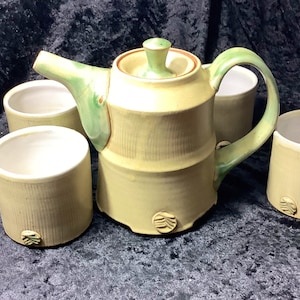 Puede incluir: Un juego de cuatro tazas de té de cerámica y una tetera con un diseño de bambú. La tetera y las tazas son de color amarillo claro con acentos verdes. Las tazas tienen un interior blanco.