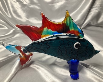 May include: A colorful glass fish sculpture with a blue tail, red and yellow fins, and a dark blue body with a white eye. The fish is standing on two blue legs.