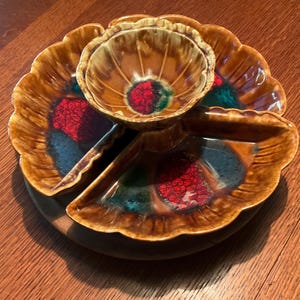 May include: A vintage ceramic serving dish with a brown, red, and teal color scheme. The dish has a central bowl and three petal-shaped sections. The dish sits on a wooden base, perfect for serving snacks or appetizers.