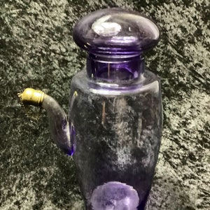 May include: A clear glass bottle with a purple hue and a gold spout. The bottle has a round, purple lid.