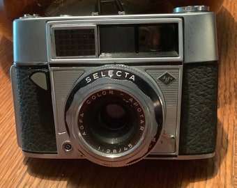 May include: Vintage silver and black Agfa Selecta camera. The camera has a black leatherette grip, a silver lens, and the text "SELECTA COLOR-APOTAR 1:2.8/45". The camera is on a wooden surface.